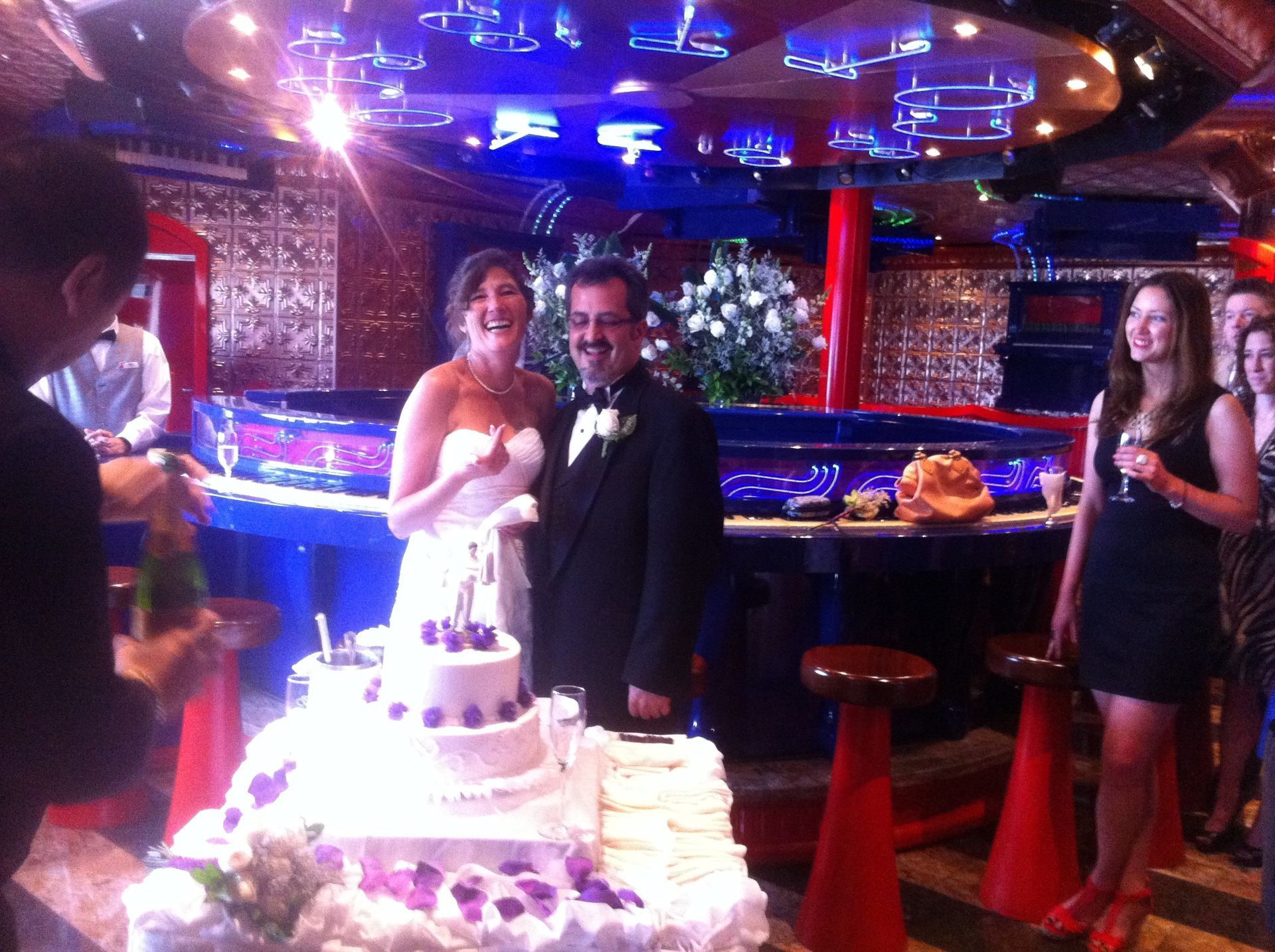Bride and groom beside a wedding cake in a neon-lit reception hall