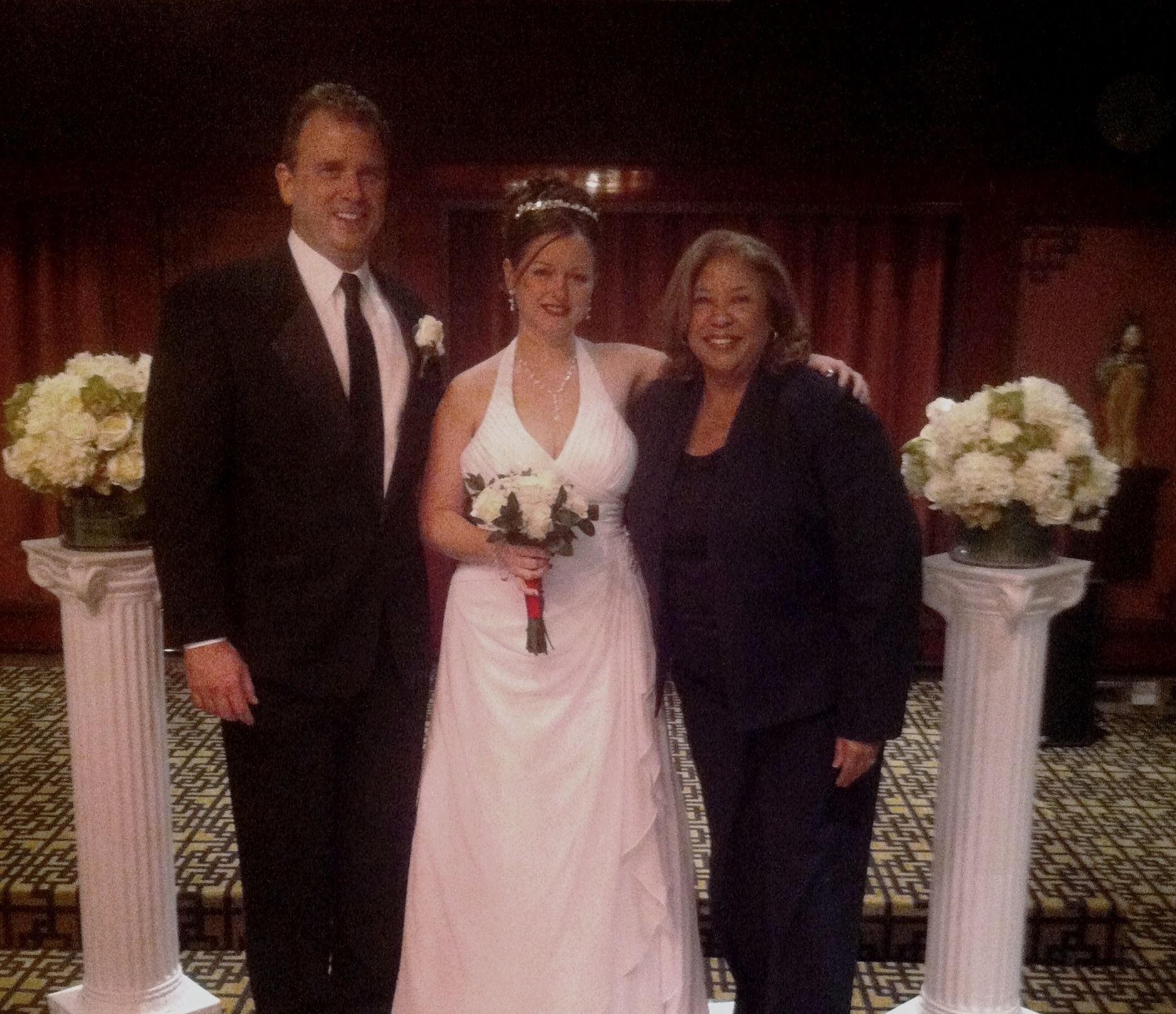 Three people pose at a wedding, with the bride in a white gown holding a bouquet.