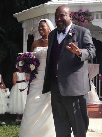 Wedding couple walking outdoors, smiling; bride holds purple bouquet, groom waves in dark suit