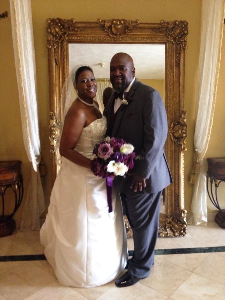 Bride and groom posing indoors before an ornate gold-framed mirror, holding a purple-and-white bouquet