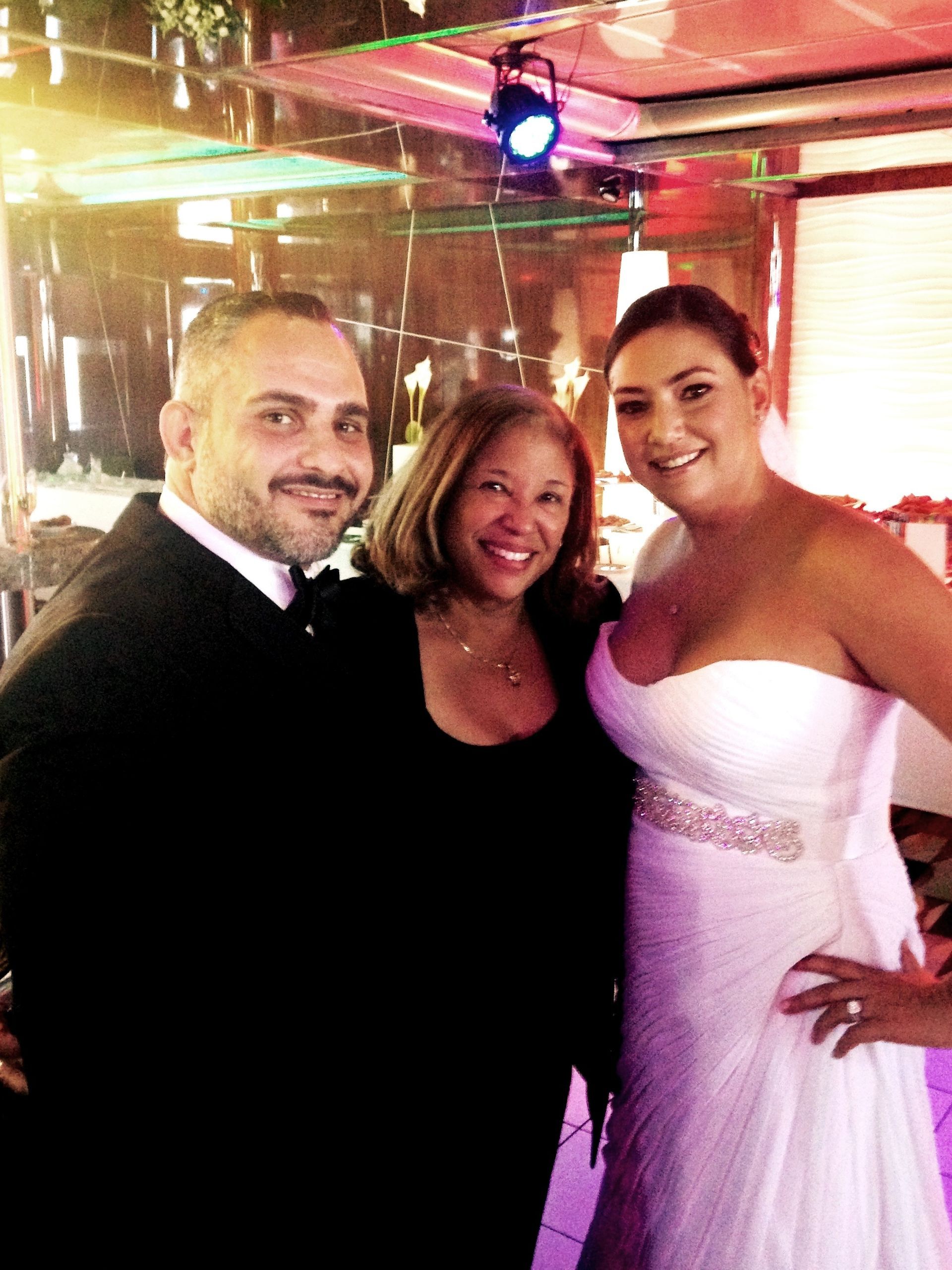 Three people smiling at a formal event, with a woman in a white dress beside two guests.