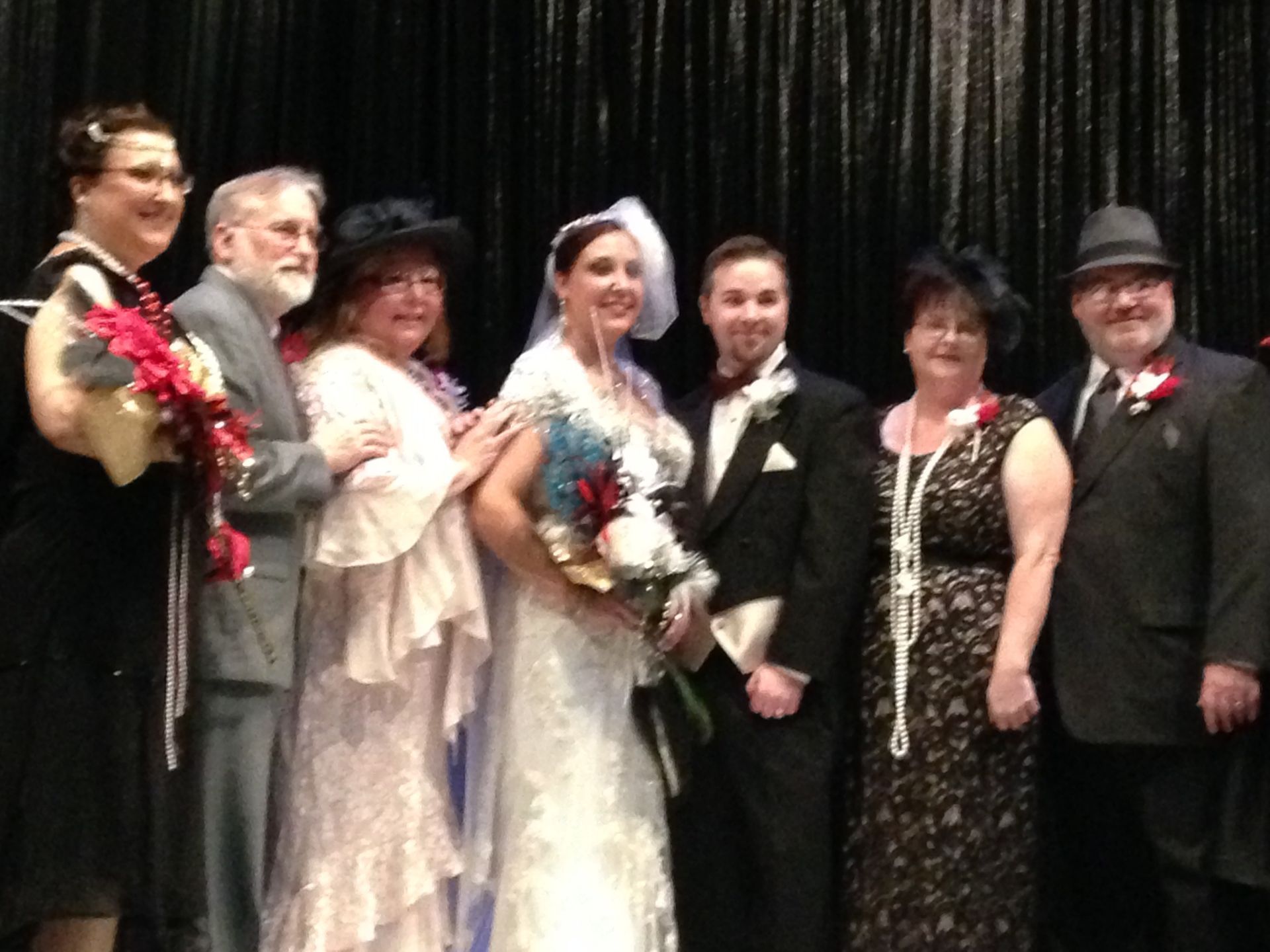 Seven people in formal attire posing on a stage in front of a black curtain.