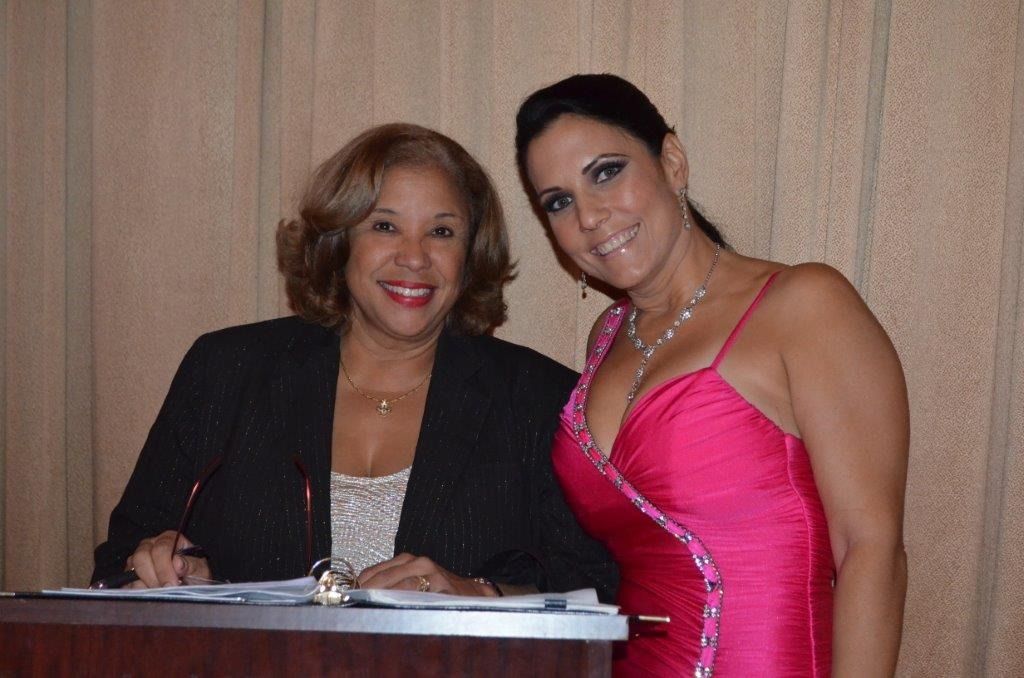 Two women posing beside a podium, one in a black jacket and one in a bright pink dress, against a wood-paneled wall