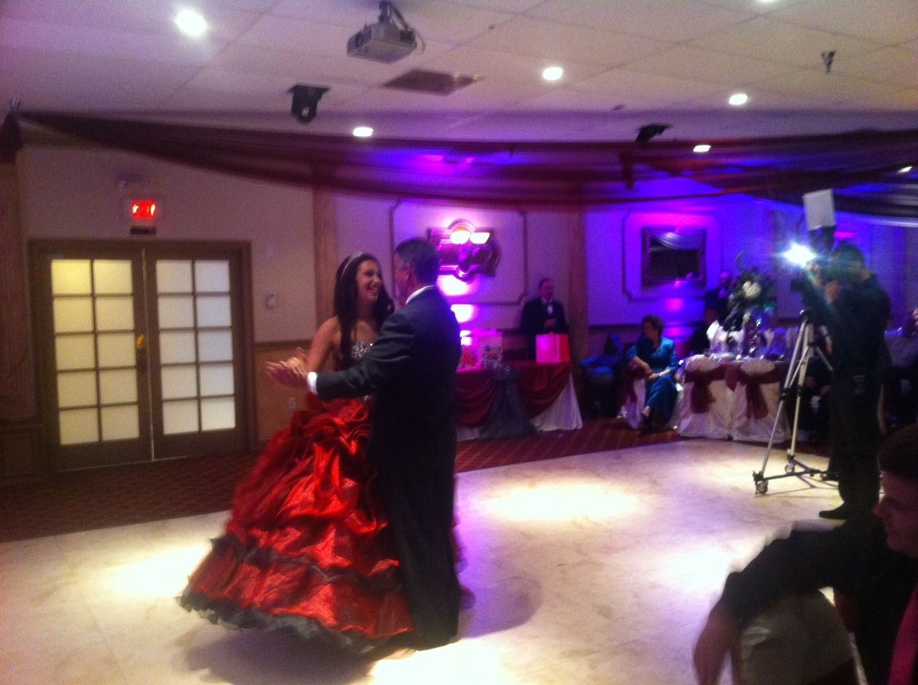 Couple dancing in a banquet hall, woman in a red gown and man in a dark suit under purple lighting.