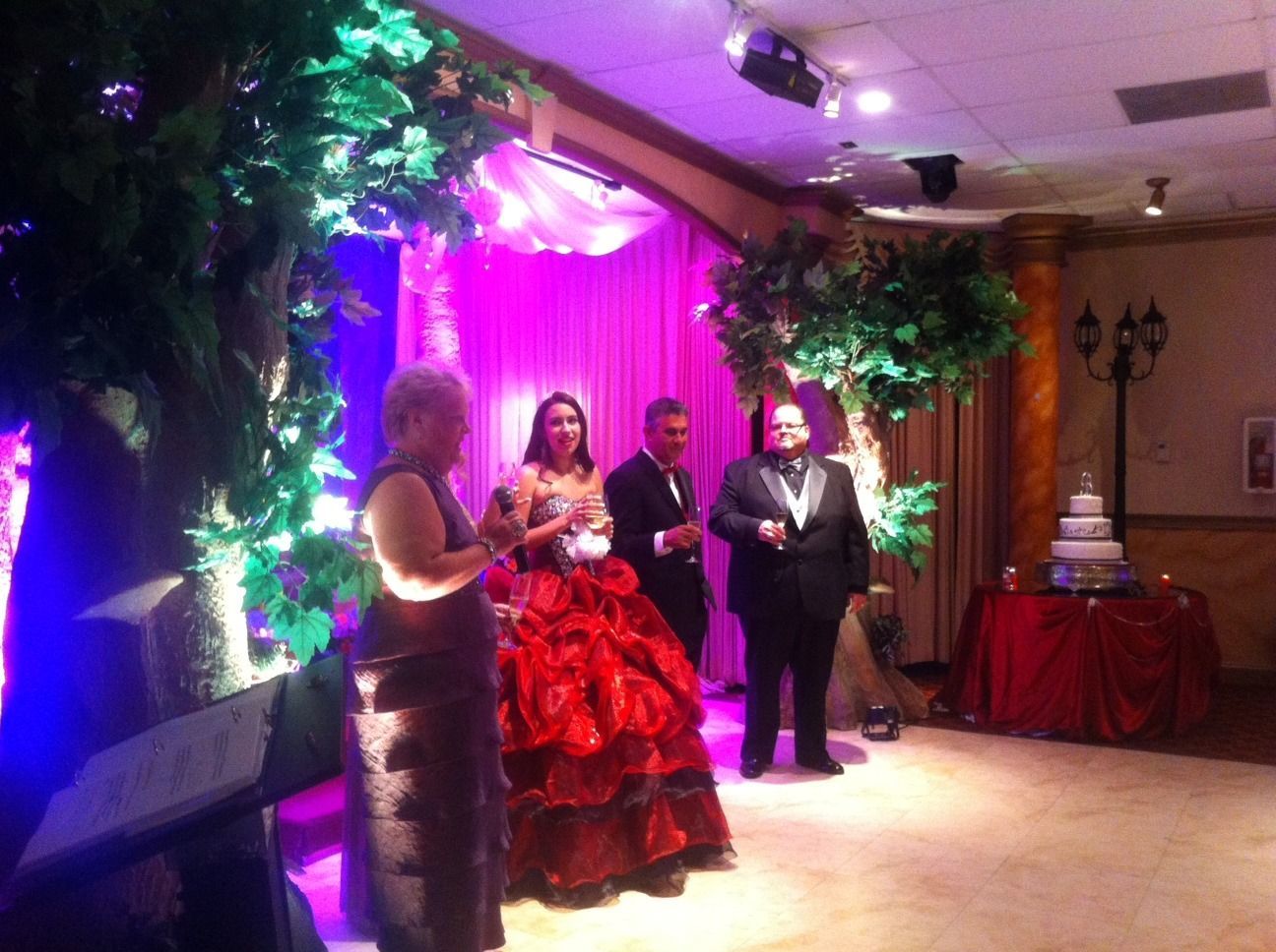 Formal event with three people on a decorated stage, purple lights, red gown, and a wedding cake nearby