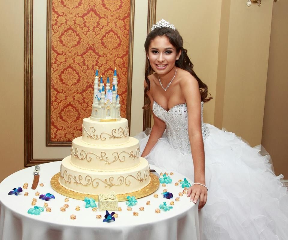 Bride in white gown beside a tiered wedding cake on a decorated table, smiling in a reception room