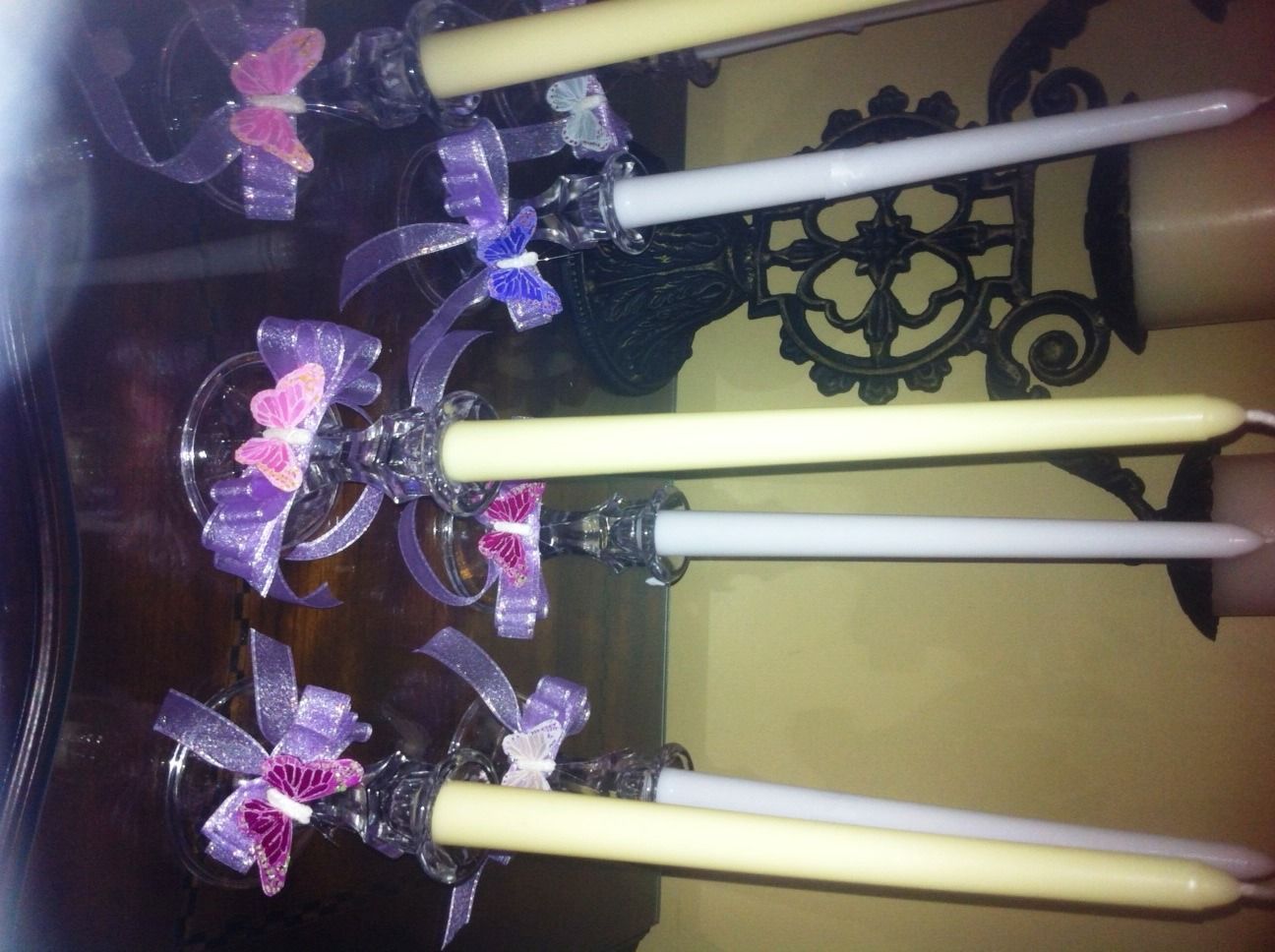 Purple floral candle holders with tall white candles on a decorative shelf, beside a wall ornament.