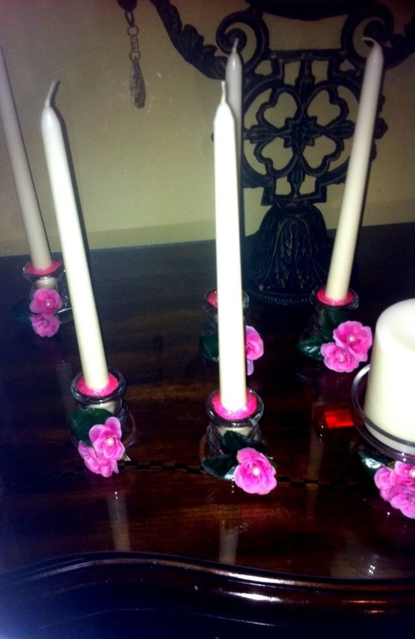 Decorated white candles in pink flower holders on a dark table with a decorative chair behind them