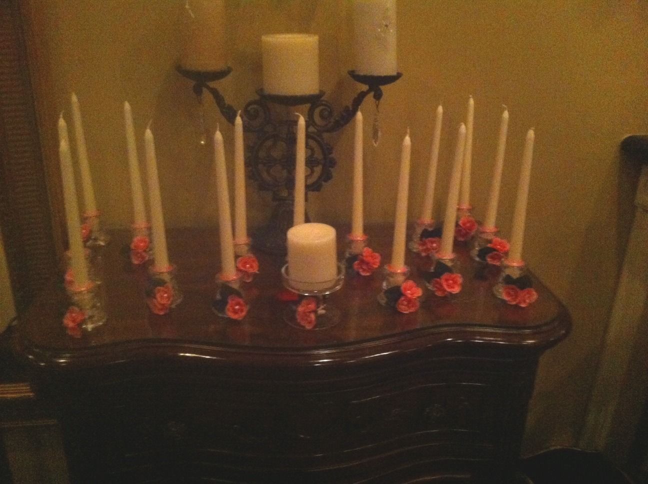 Six white candles and red flowers arranged on a dark wooden dresser against a beige wall.