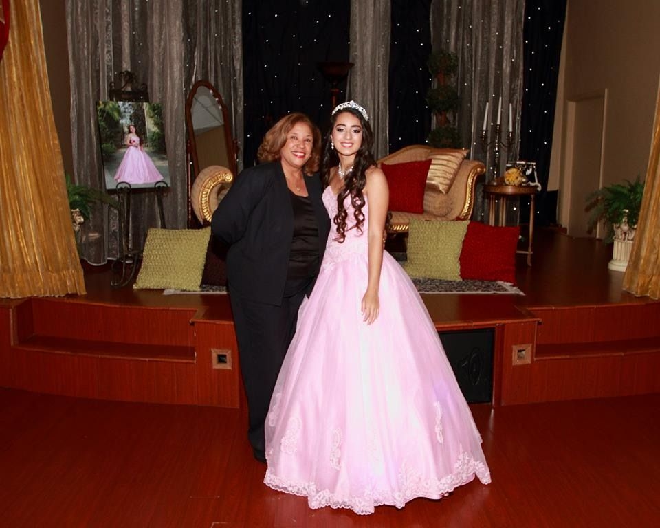 Two people posing indoors, one in a pink formal gown and tiara, beside a stage with red curtains.