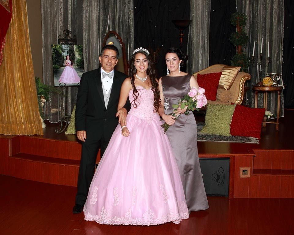 Three people posing on a stage, with one in a pink gown and two in formal attire, holding flowers.