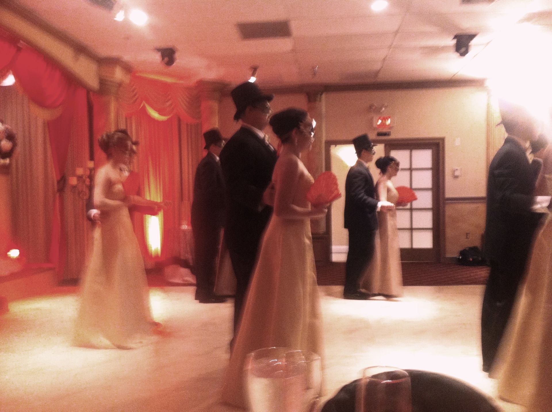 People in formal wear standing in a softly lit banquet hall with red drapes and a dance floor