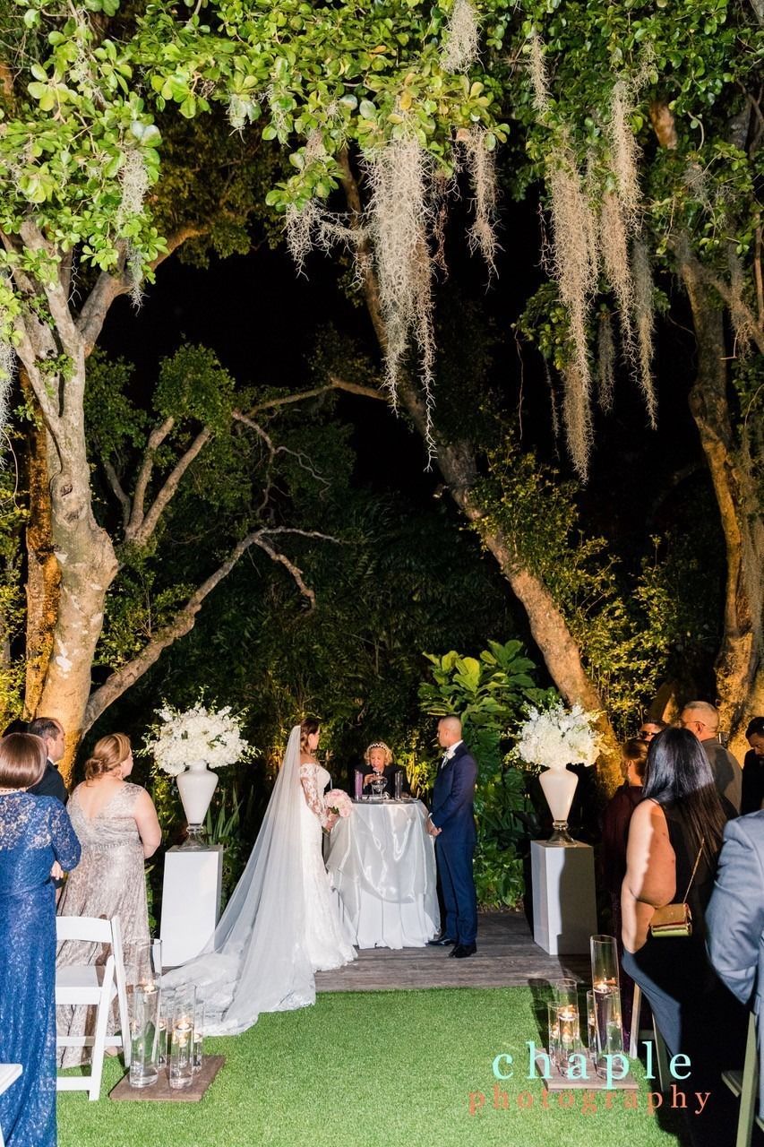 Outdoor nighttime wedding ceremony beneath large trees, with bride and groom at the altar and guests seated nearby