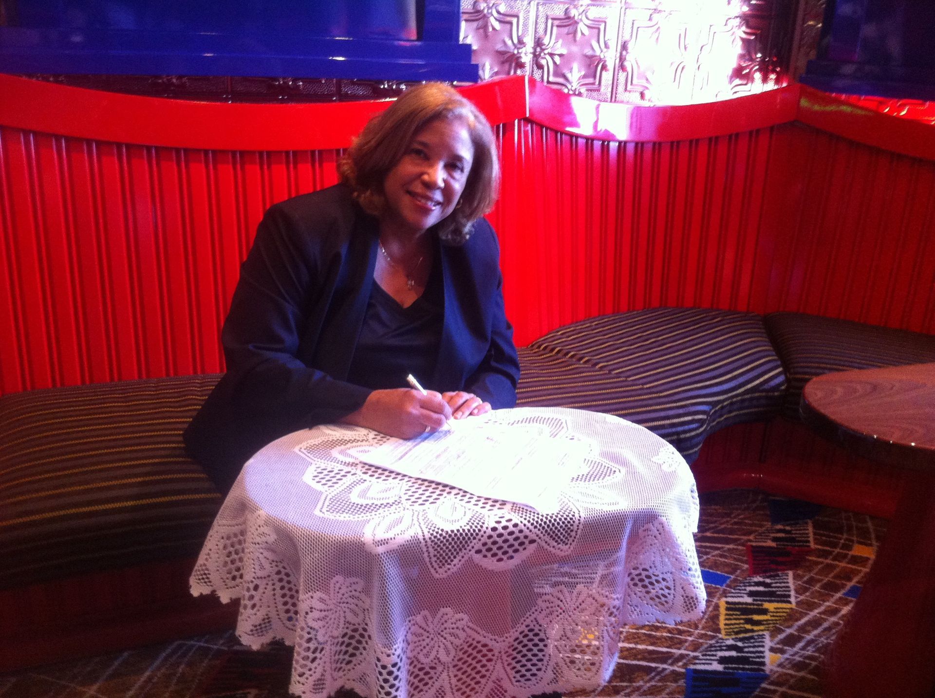 Person seated at a lace-covered table in a red-lit lounge, smiling while writing on paper
