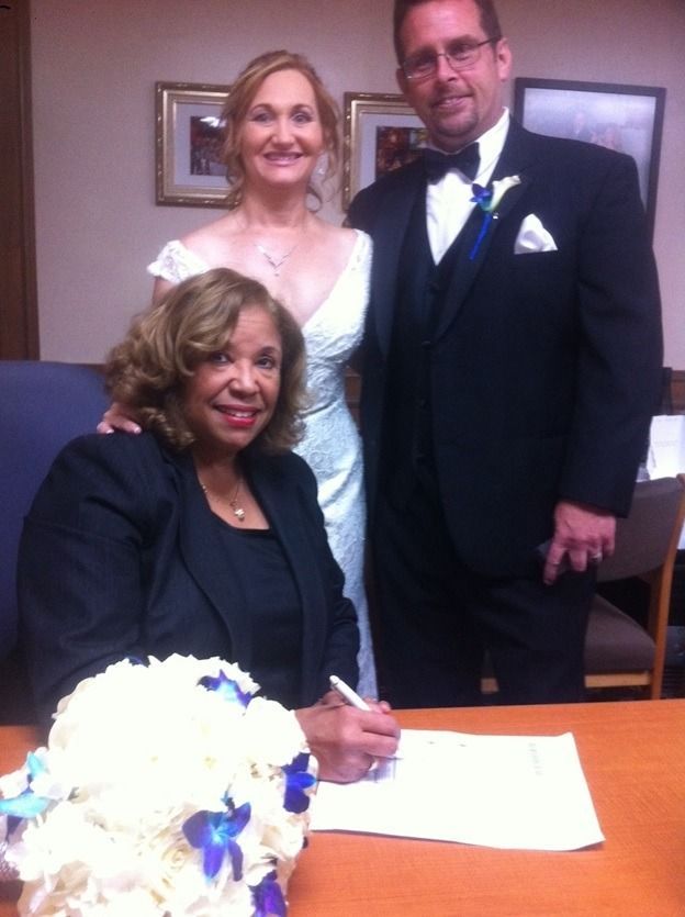 Woman in black signing a document, with a bride and groom standing behind her at a wedding table.