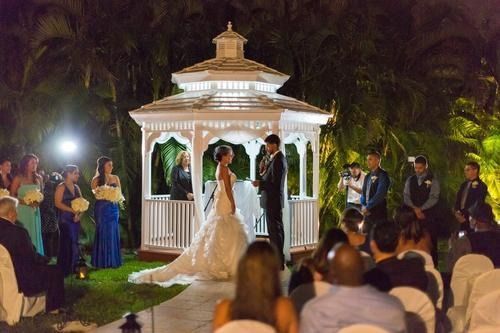 Outdoor wedding ceremony in a lit gazebo at night, with bride, groom, and guests gathered around.