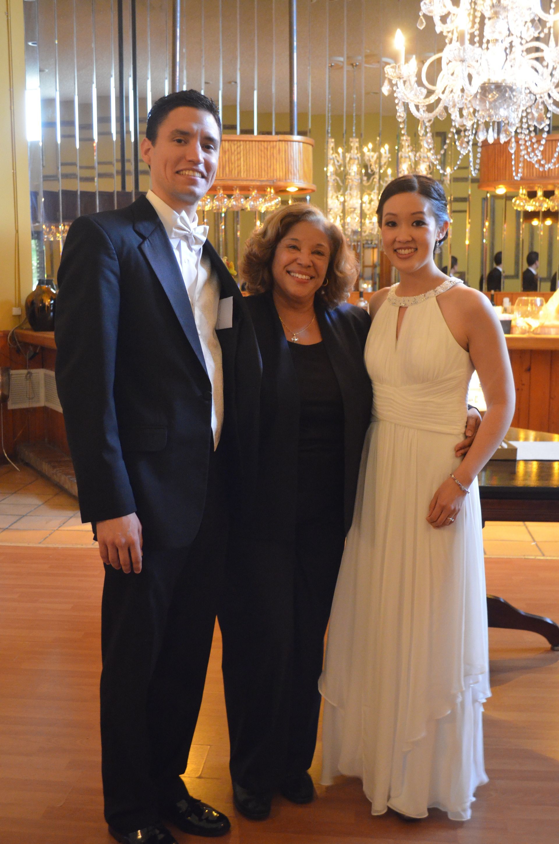 Three people posing together in formal attire inside a chandelier-lit ballroom.