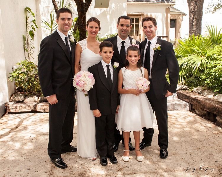 Wedding party posing outdoors in formal attire, with bride, groom, and children holding flowers.