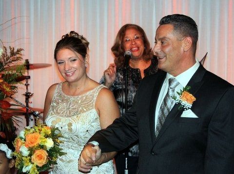 Bride and groom smiling and holding hands at their wedding reception, with a singer in the background