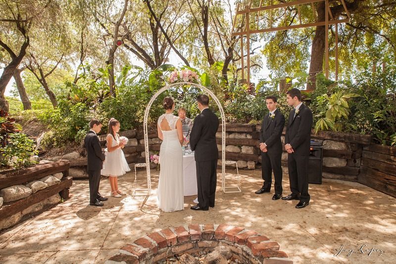Wedding ceremony outdoors under trees, with bride and groom at the altar and attendants nearby.
