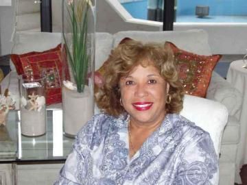 Woman smiling on a white couch in a bright living room with patterned pillows and a glass table.