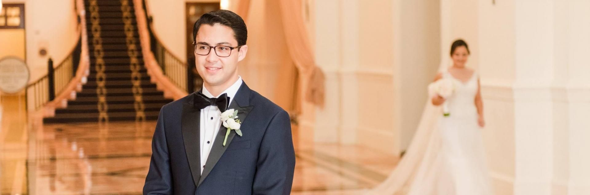 Groom in a navy suit stands in a grand hall; bride in a white gown is softly blurred in the background.