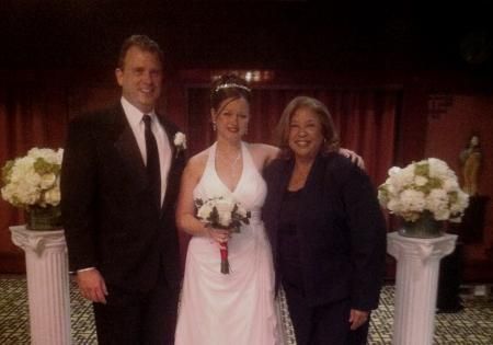 Three people pose at a wedding, with the bride holding a bouquet between two guests.
