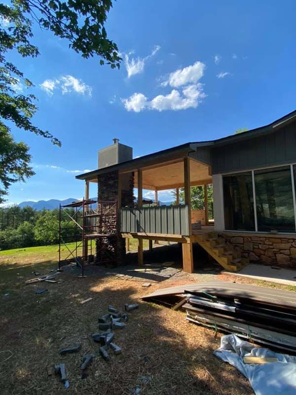 House with deck under construction on a sunny day. Brick chimney, mountain views, and lumber on the ground.