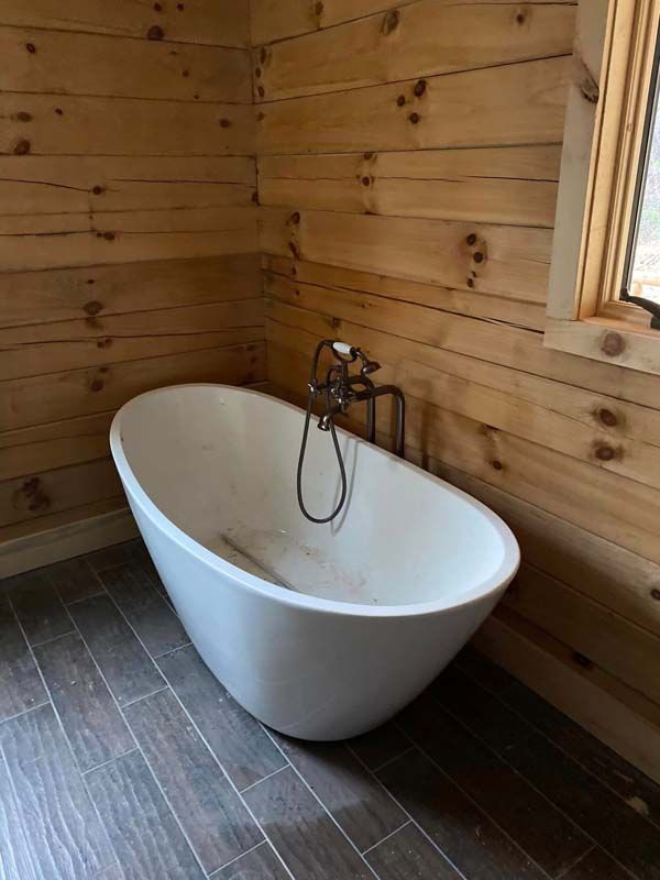 Freestanding white bathtub in a wood-paneled bathroom with gray tile floor, next to a window.