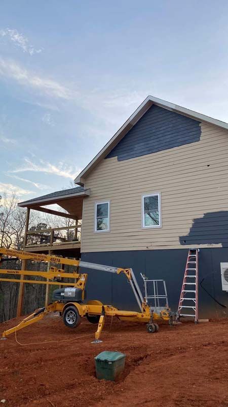 Yellow lift machinery next to a house with beige and blue siding, a porch, and a ladder.