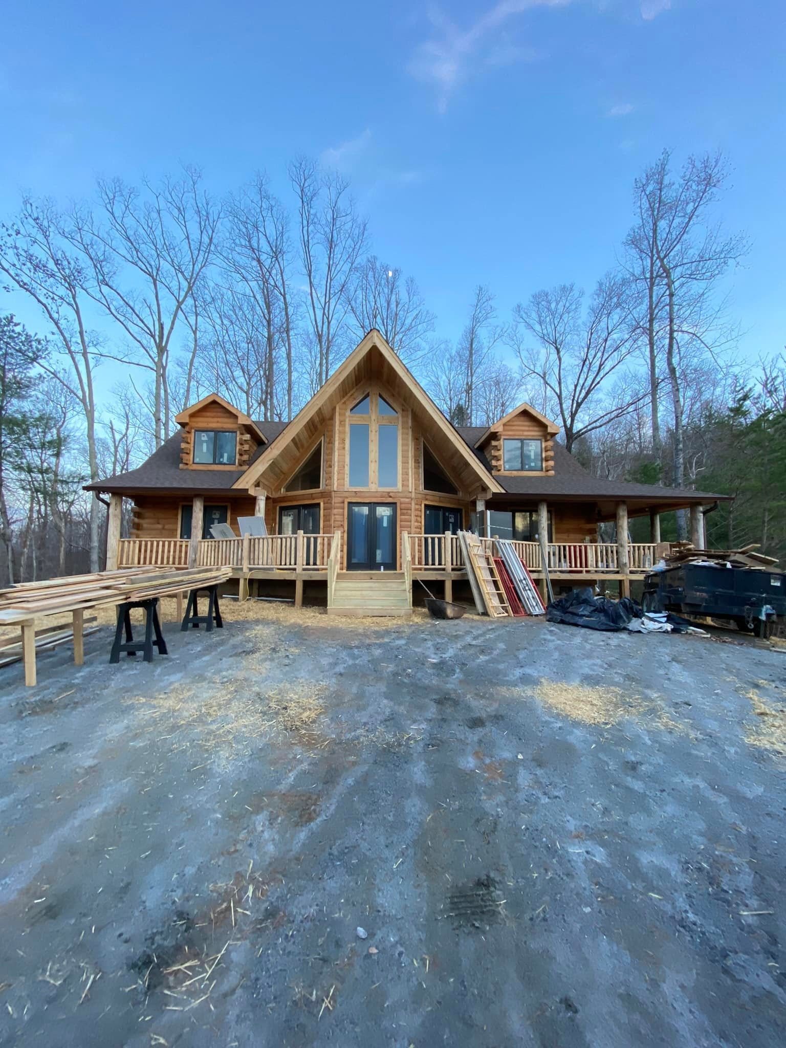 Log cabin under construction with a large front deck, surrounded by trees and a blue sky.