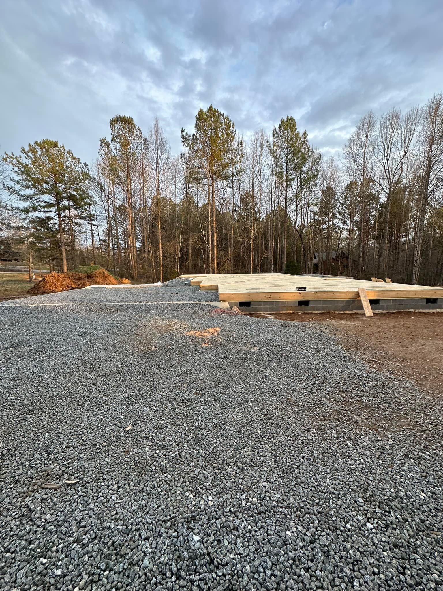 Gravel pile in foreground; wood foundation and trees in background under cloudy sky.