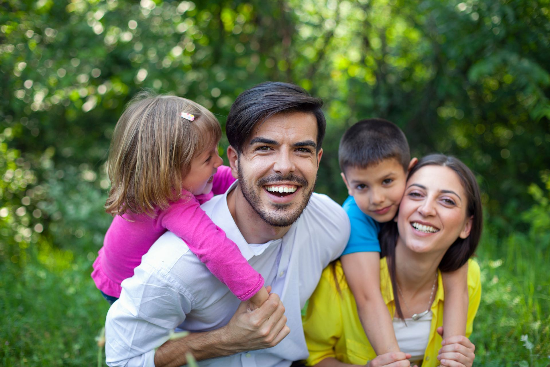 Family smiling together outdoors in a green park, with two adults and two children hugging.