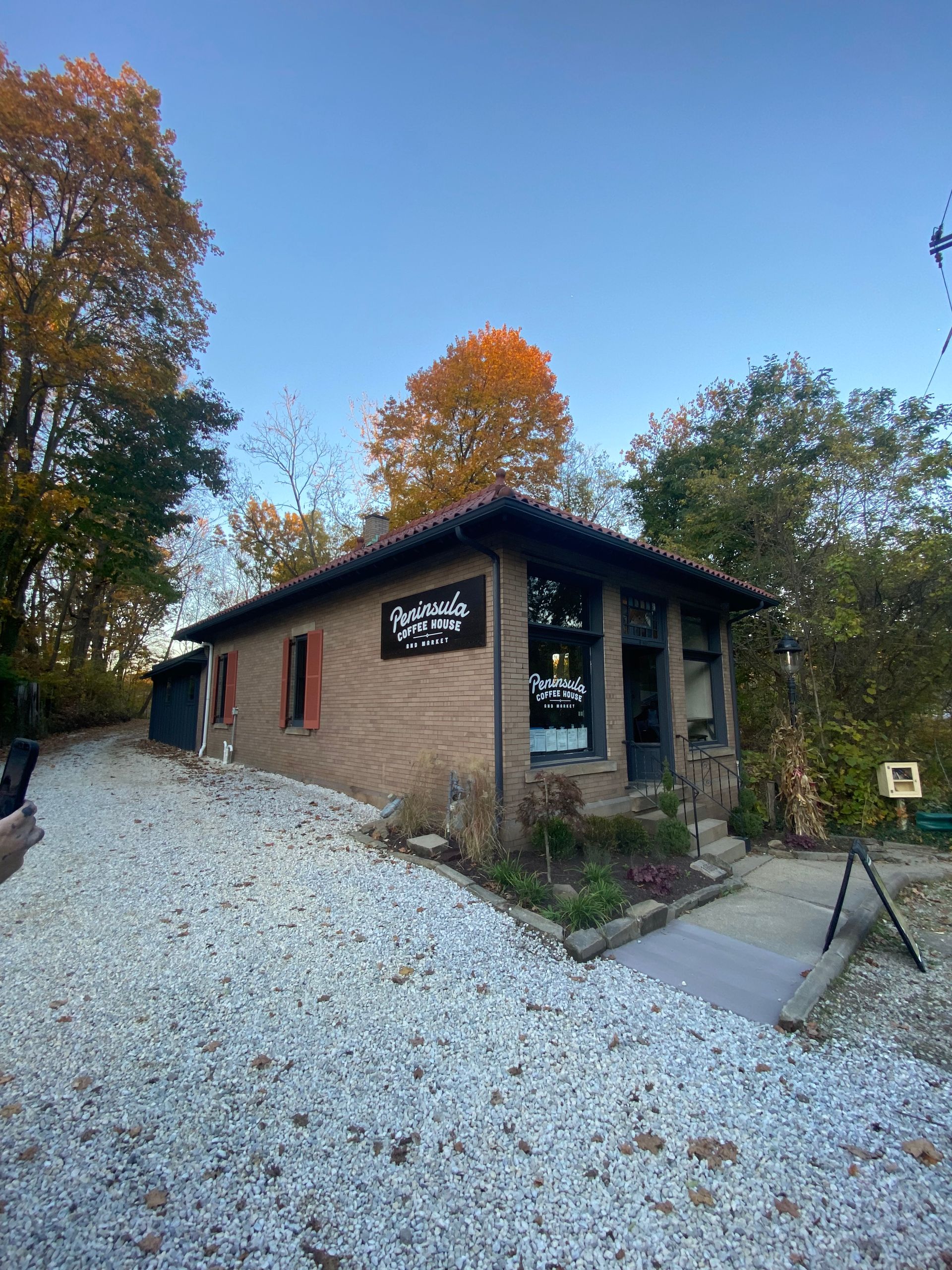 Small brick building with signage, gravel driveway, surrounded by trees under a clear blue sky.
