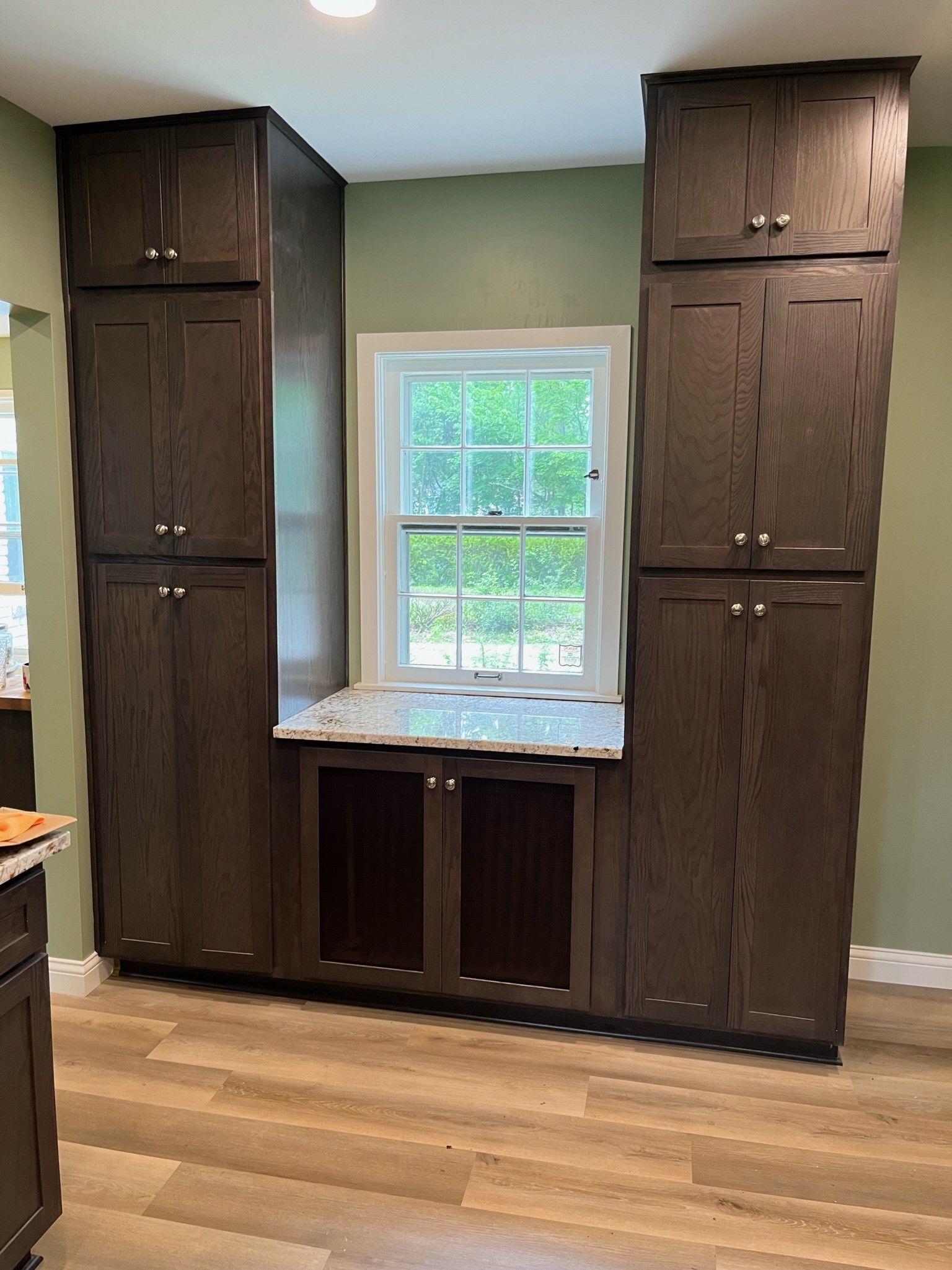 Dark brown kitchen cabinets flanking a window with a granite countertop; green wall; light wood floor.