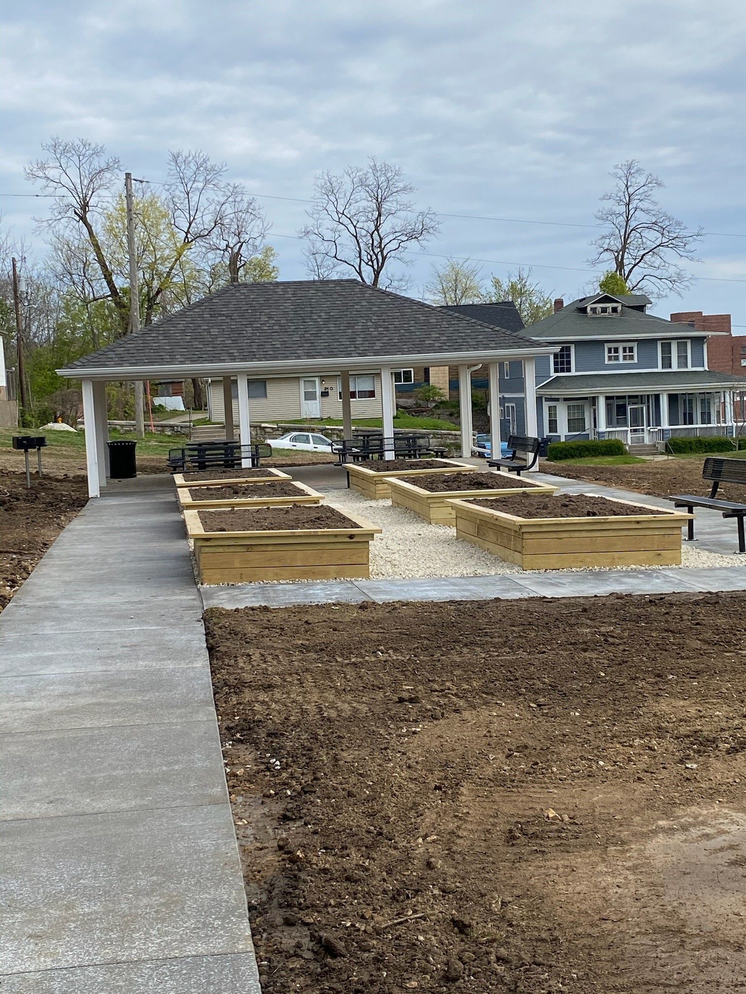 Raised garden beds under a white-roofed gazebo; concrete path; bare trees and a house in background.