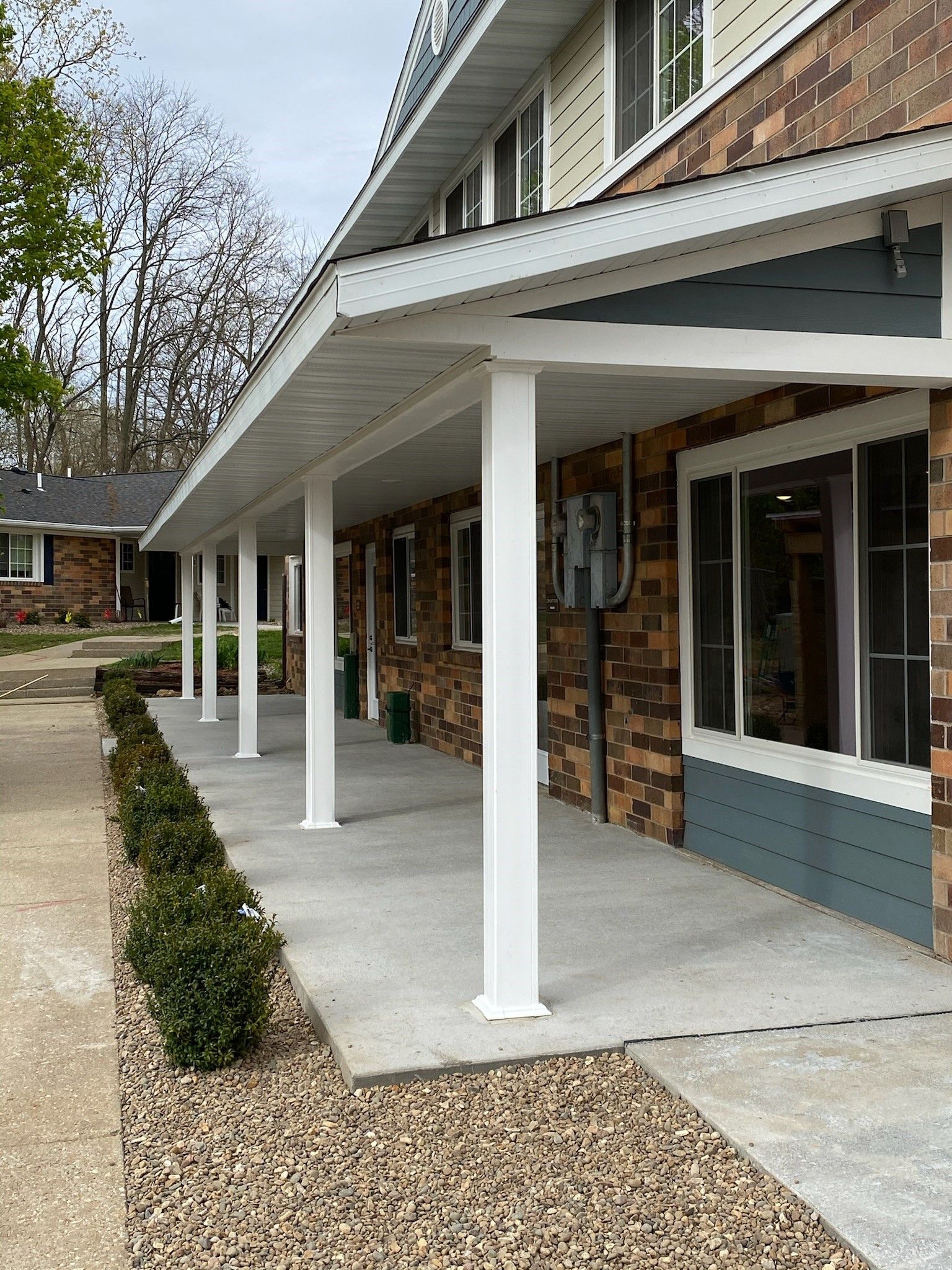 White-columned porch with brick and teal siding. Low bushes line the edge. Gray concrete and gravel walkway.