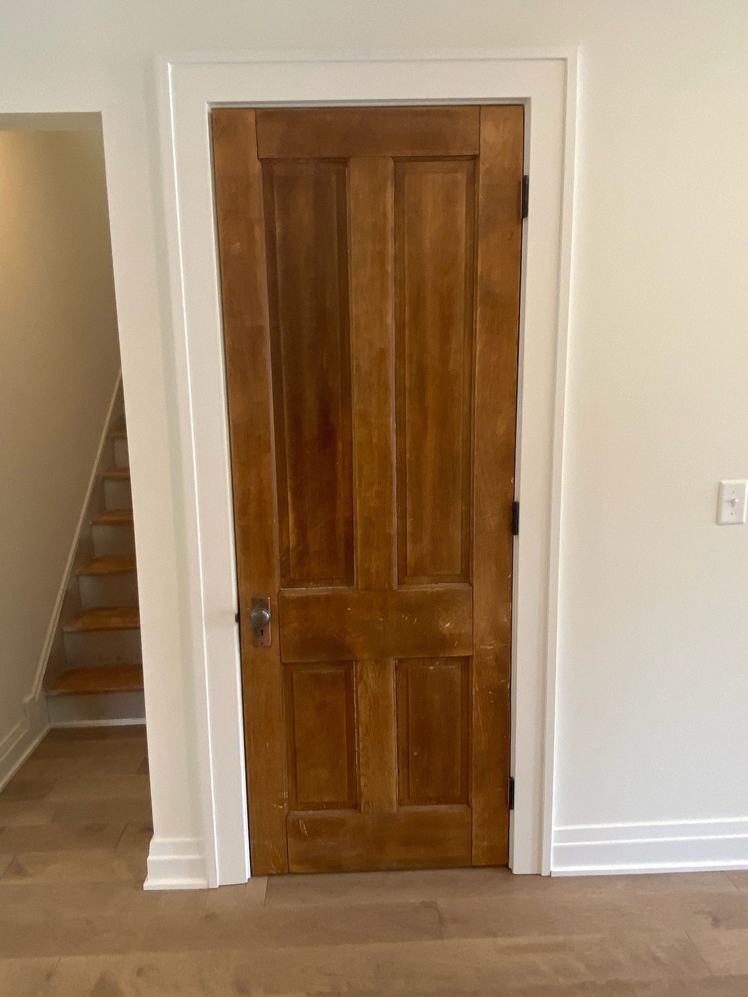 Wooden door with white trim in a room with stairs on the left and wood floors.