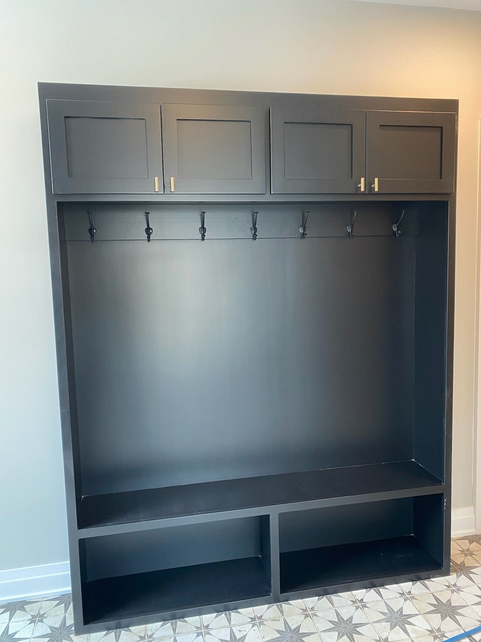Black built-in mudroom with cabinets, hooks, bench, and lower storage shelves against a light gray wall.