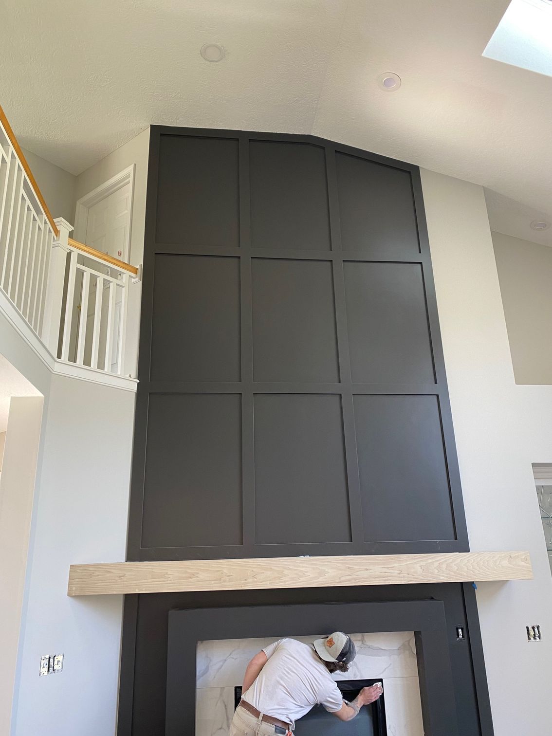 Tall charcoal-colored paneling with a fireplace and mantle, being worked on by a man in a white hat.
