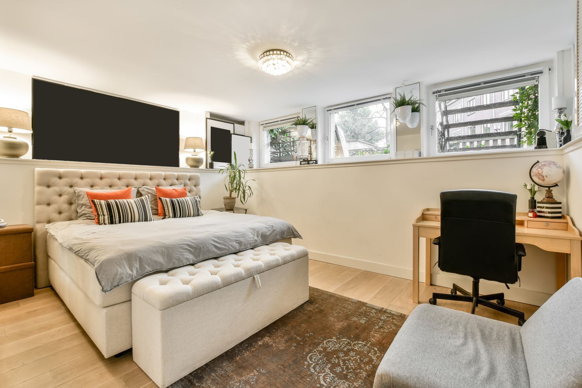 Bedroom with bed, desk, TV, and windows. Neutral colors, wood floors, and decorative pillows.