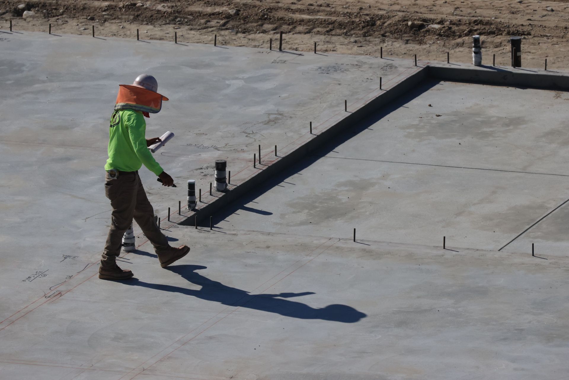 Worker in green safety vest and orange hard hat walking on a concrete slab at a construction site