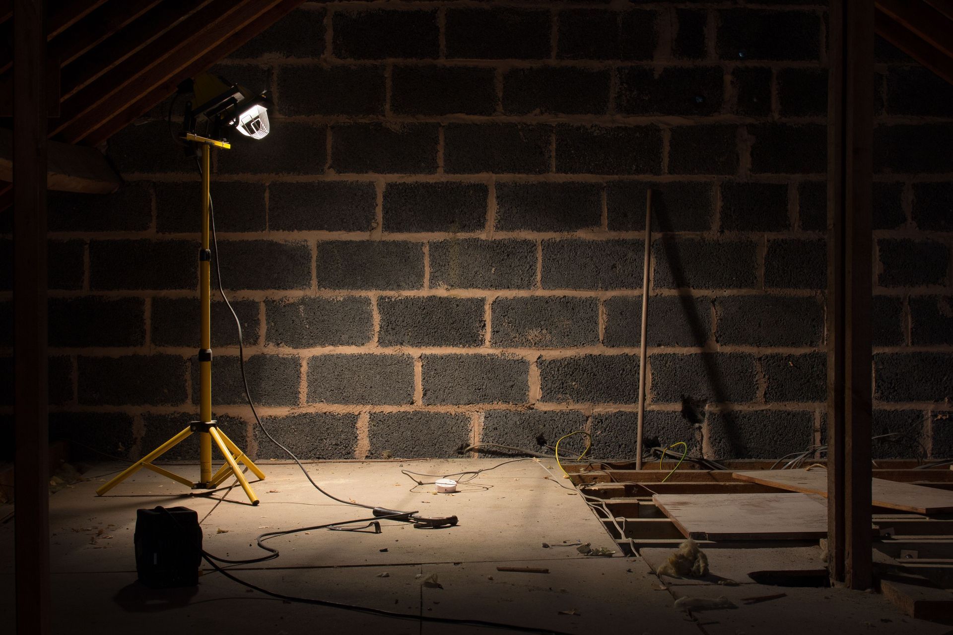 Dim construction site with a yellow tripod work light illuminating a brick wall and scattered tools.