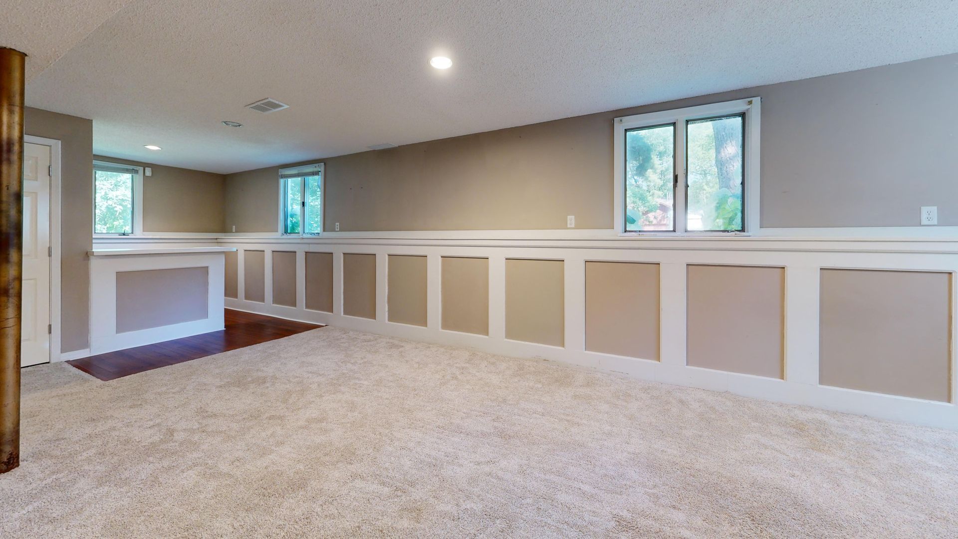 Spacious carpeted room with white wainscoting, beige walls, and small windows letting in natural light