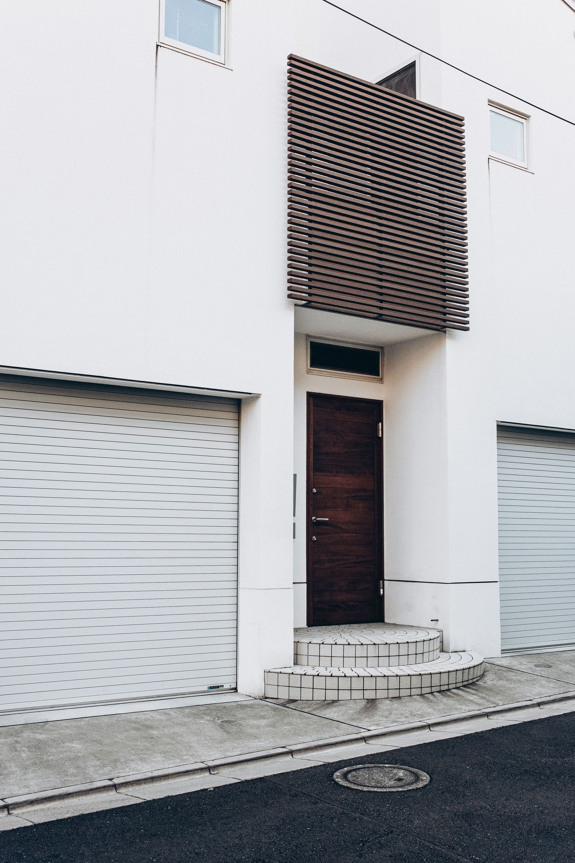 Modern white building facade with a brown door, roll-up garage doors, and a decorative brick screen above the entrance
