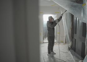 Person in protective suit spraying white coating on a room ceiling during renovation work