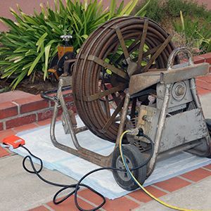 Technician kneels on tiled floor, repairing a drain near a spinning floor machine and red extension cord.