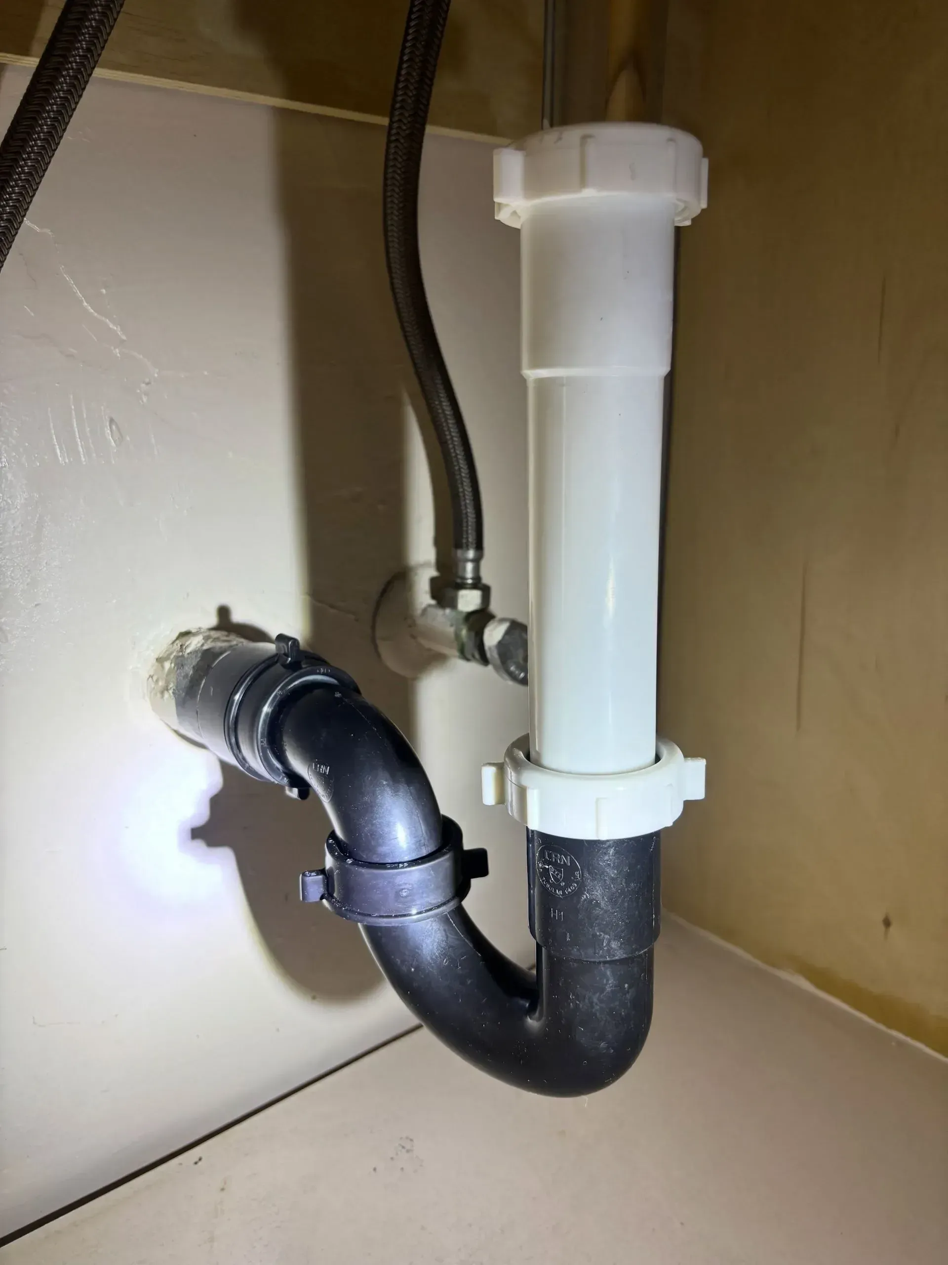 Under-sink plumbing with black P-trap and white drain pipe against a beige wall