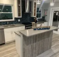 Modern kitchen with white cabinets, blue backsplash, and light wood flooring. Island with a gray countertop.