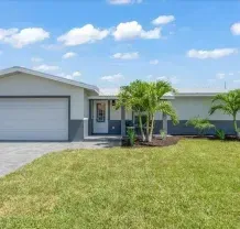 A single-story house with a grey garage door, white walls, and a green lawn under a blue sky.